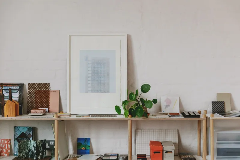 Timber shelf displaying framed artwork, material samples, and a potted plant against a textured plaster wall