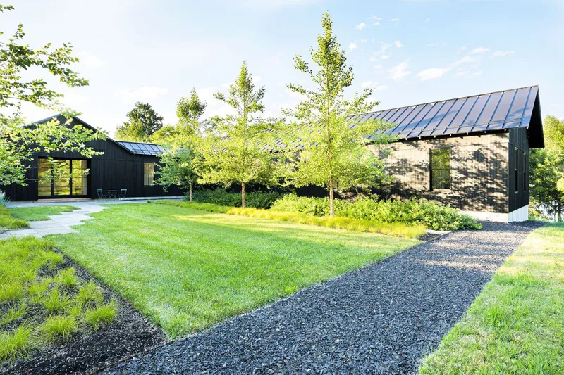 Gravel path leading to two black-clad gable volumes separated by young trees in a meadow setting