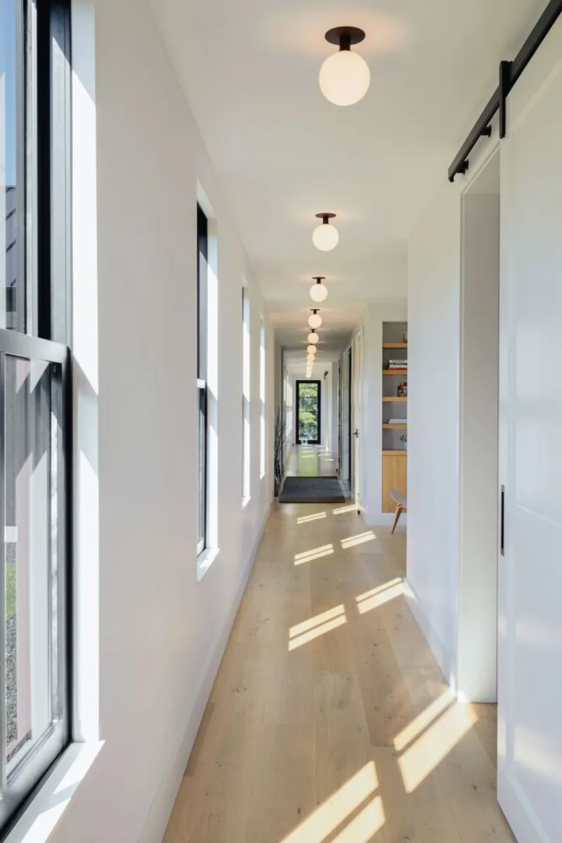 White hallway with globe ceiling fixtures and rectangular windows casting striped shadows on pale wood flooring