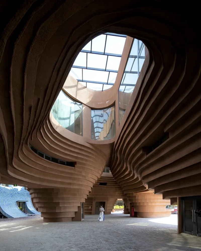 Layered timber ceiling forms spiraling above a ground-floor lobby with a figure in white passing through