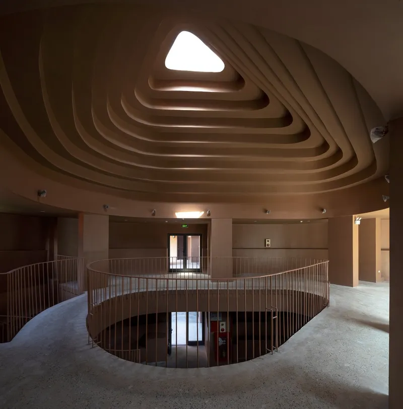 Interior balcony with curved metal railing beneath a triangular layered ceiling opening illuminated by daylight