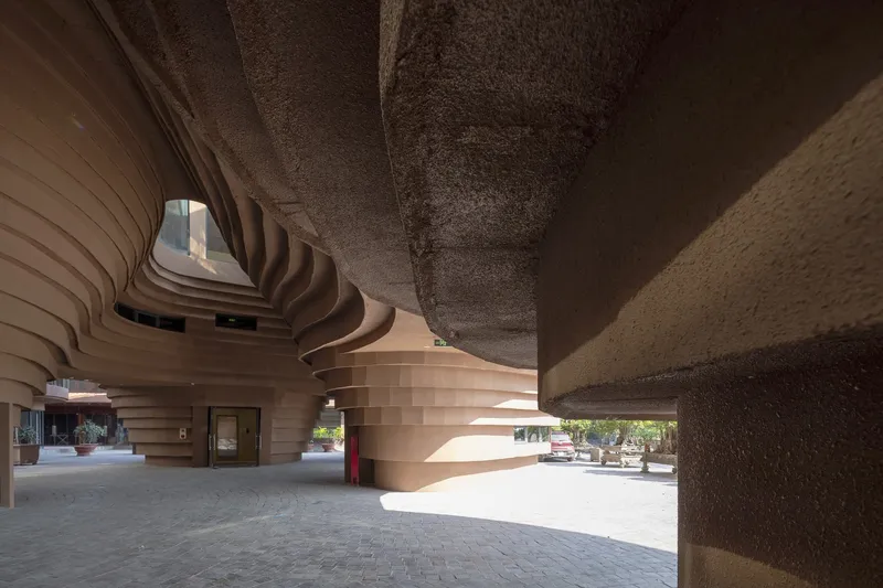 Ground level passageway beneath textured concrete slabs and horizontally striped curving walls in afternoon light