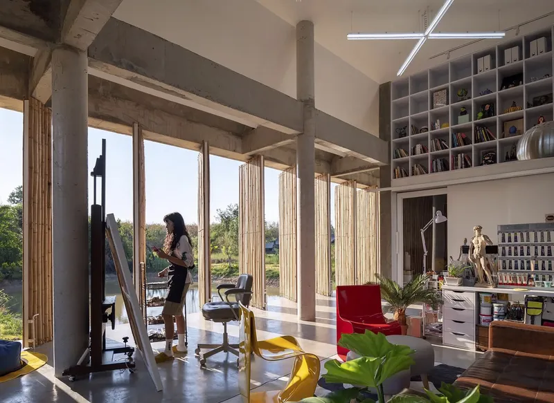Interior workspace with floor-to-ceiling bamboo screens, concrete beams and a person painting at an easel