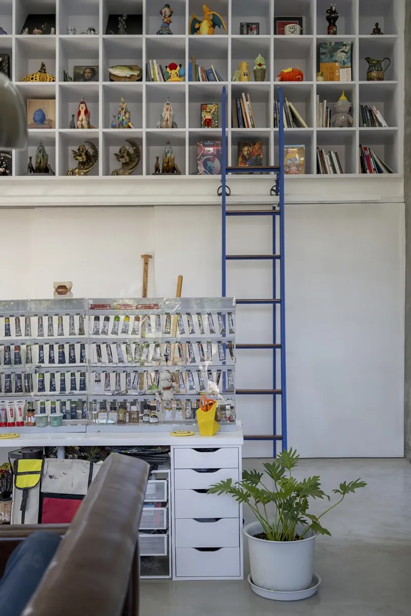 White shelving wall filled with figurines and books above a paint-stocked workspace with a blue rolling ladder
