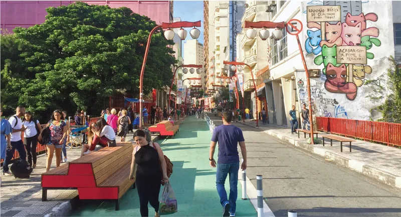 Pedestrianised street with red benches, green paving and mural-clad buildings under a large tree