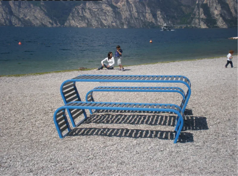 Pair of undulating blue metal benches on a gravel beach with families near the waterline