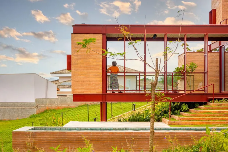 Elevated red steel frame pavilion with glazed walls overlooking a lawn with young trees