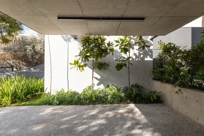 Covered entry drive with exposed concrete soffit, white walls, and planted garden bed in dappled shade