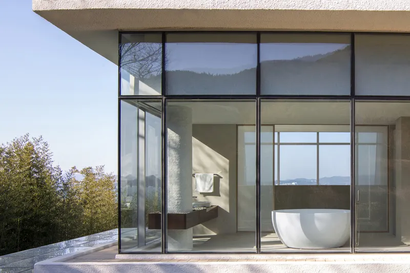 Corner glazing framing a freestanding white bathtub with hillside views beyond the terrace