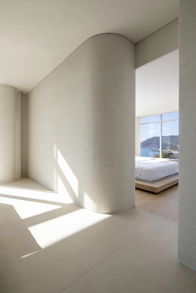 Cylindrical plaster column catching sunlight in a corridor leading to a bedroom with coastal views