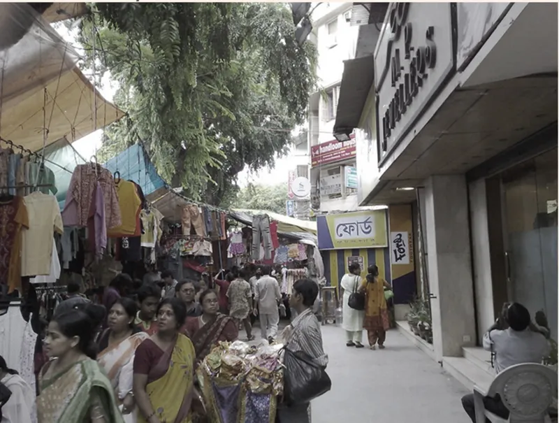Crowded market street with pedestrians among stalls and storefronts beneath hanging laundry and overhanging trees