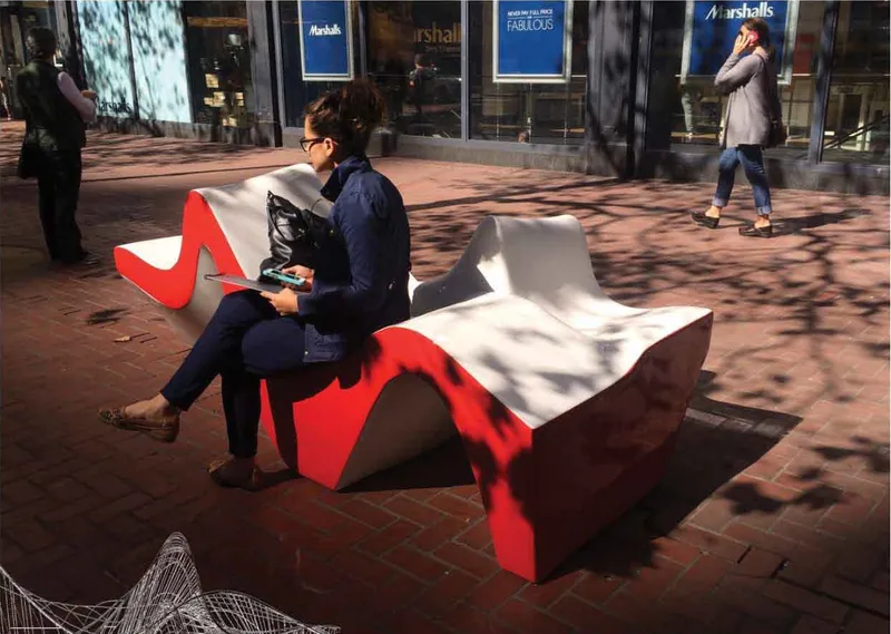 Red and white undulating public bench on brick plaza with pedestrians passing in sunlight