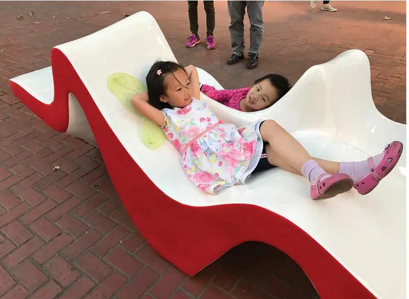 Two children reclining on the curved white seat surface of the wavy bench sculpture