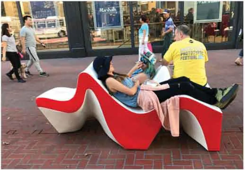 Side view of the sinuous bench with two seated figures reading beside retail storefronts