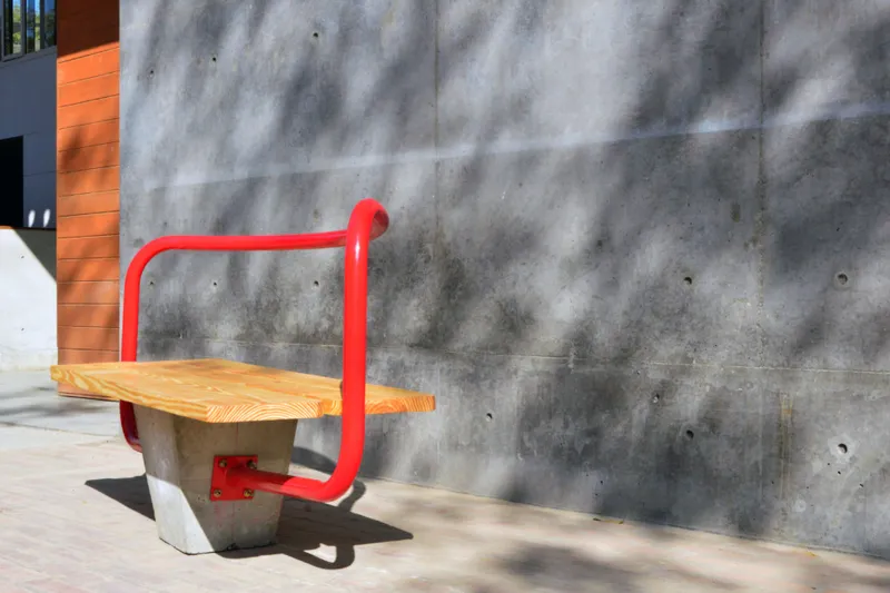 Outdoor bench with timber seat and red tubular steel frame against a weathered concrete wall