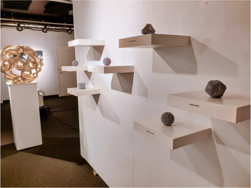 Exhibition view of geometric sculptures on tiered white pedestals under track lighting
