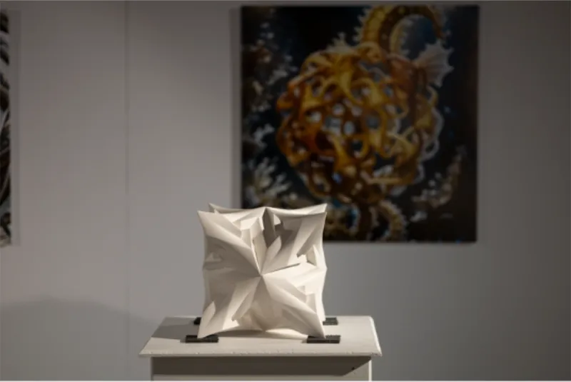 White polyhedral plaster sculpture on a pedestal against a gallery wall with artwork behind