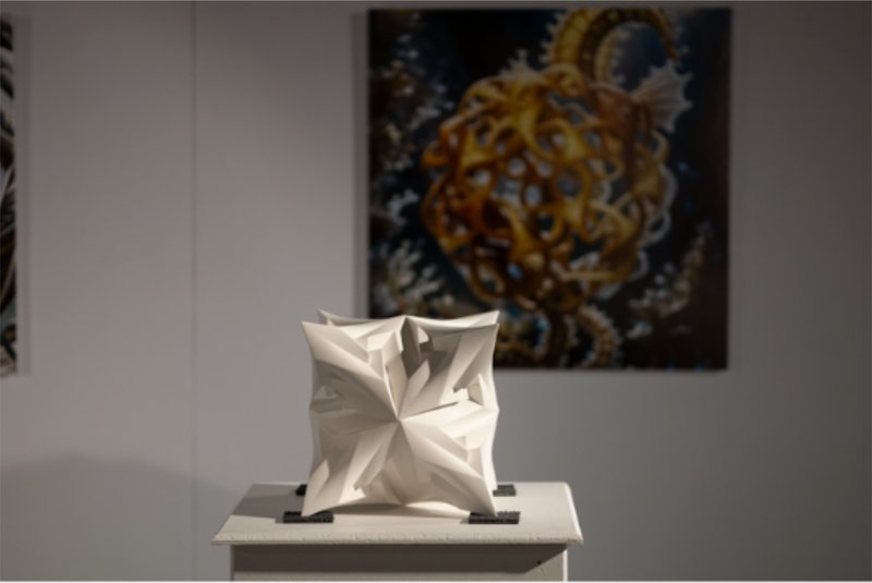 White polyhedral plaster sculpture on a pedestal against a gallery wall with artwork behind