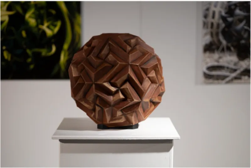 Spherical wooden sculpture with faceted geometric surface displayed on a white pedestal in gallery lighting