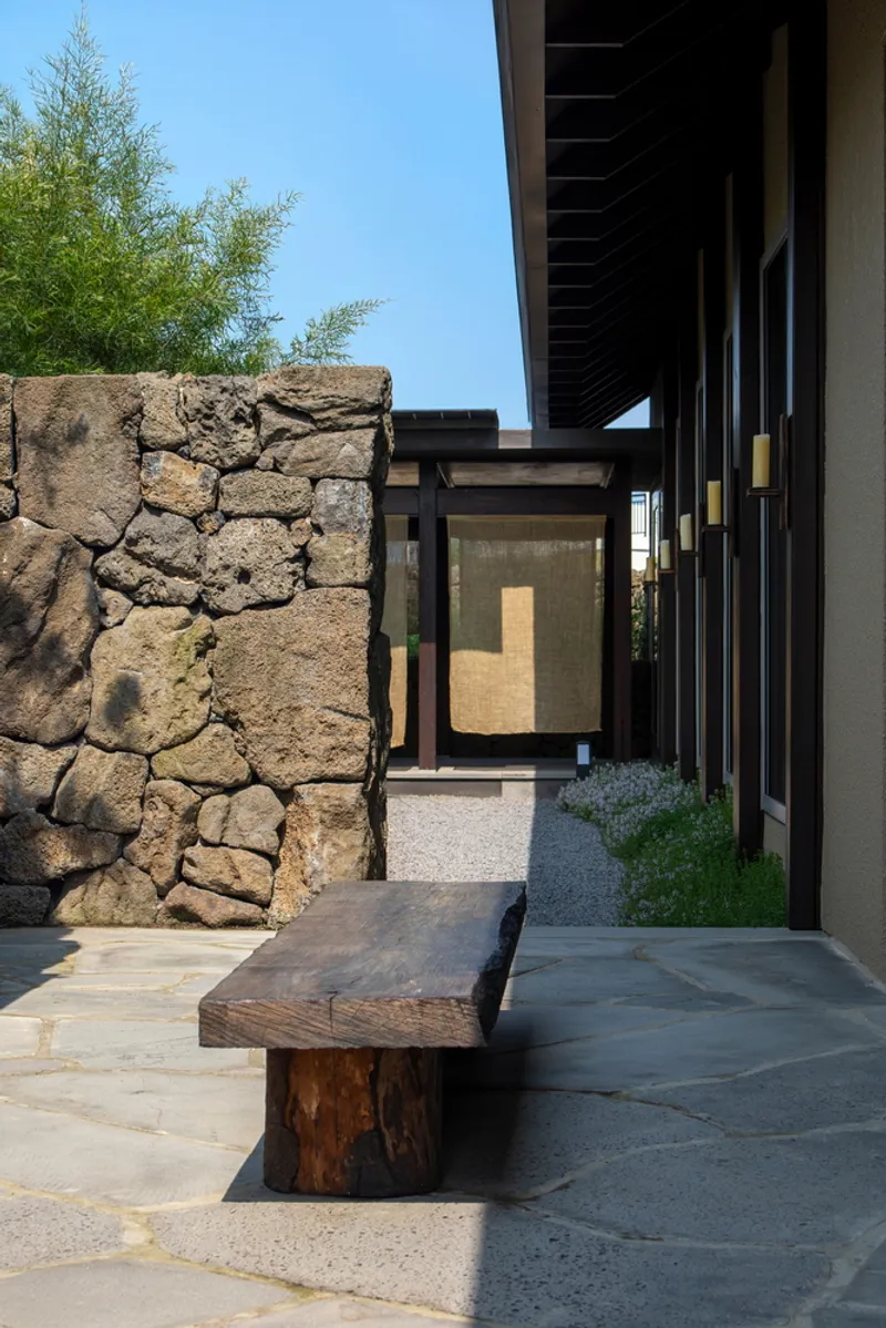 Outdoor courtyard with rough volcanic stone wall, timber bench and steel-framed glazing under clear blue sky