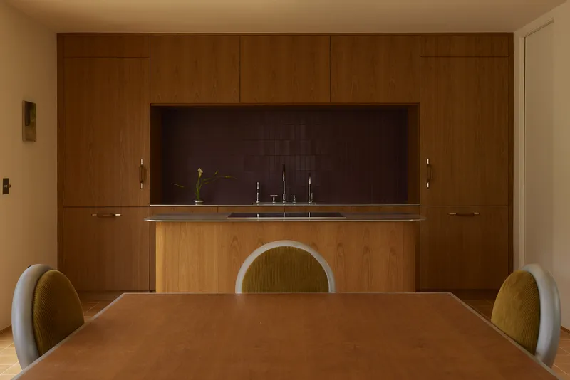 Dining table with curved-back chairs facing a wood-paneled kitchen with dark tile backsplash and stainless sink