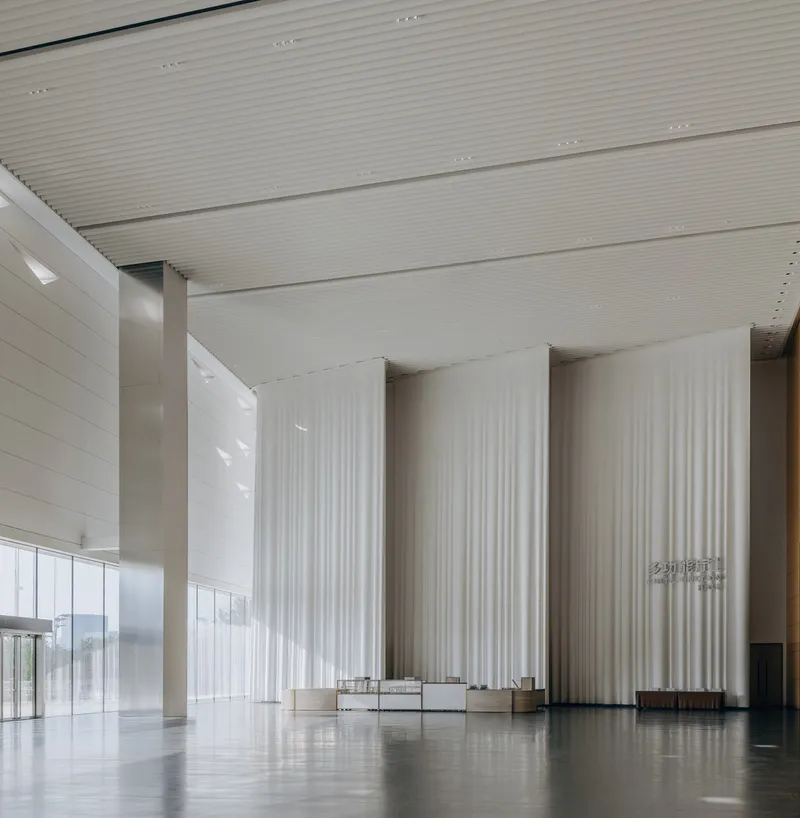 Double-height lobby with ribbed ceiling panels and floor-to-ceiling fabric curtains over stone flooring