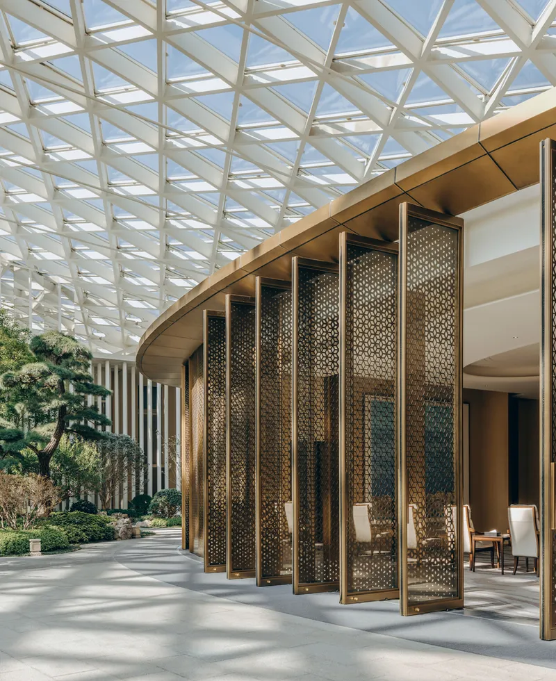 Perforated bronze folding screens framing an entrance beneath the glazed diagrid roof structure