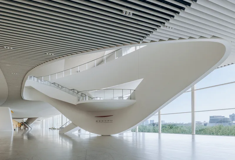 Flowing sculptural staircase in a white interior with glass curtain wall overlooking green landscape