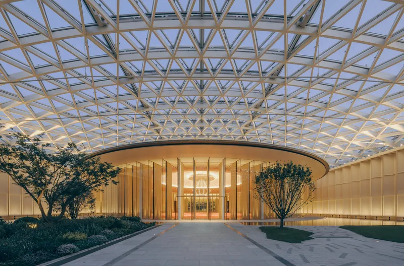 Covered entrance courtyard with triangulated glass roof and cylindrical louvered volume at dusk