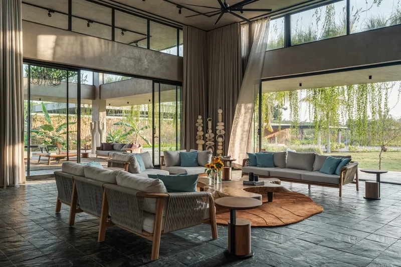Tall living room with stacked concrete beams and floor-to-ceiling curtains framing views of cascading greenery