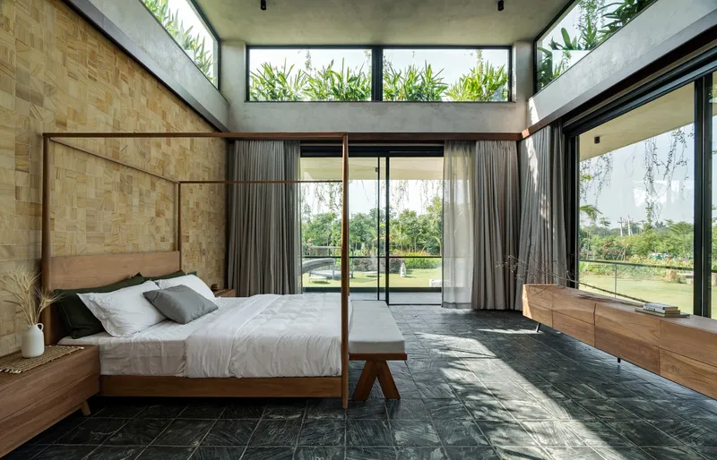 Bedroom with woven wood headboard wall and full-height glazing opening onto lawn under concrete ceiling