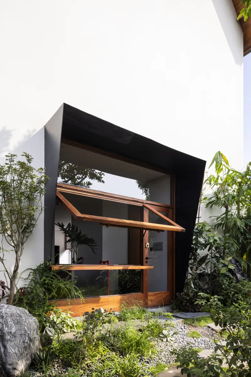 Timber-framed entrance with sloped canopy set within a black-framed aperture surrounded by planted beds