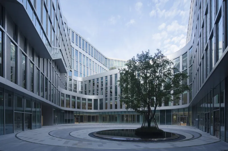 Circular courtyard with concentric paving rings around a central tree beneath curved glass facades