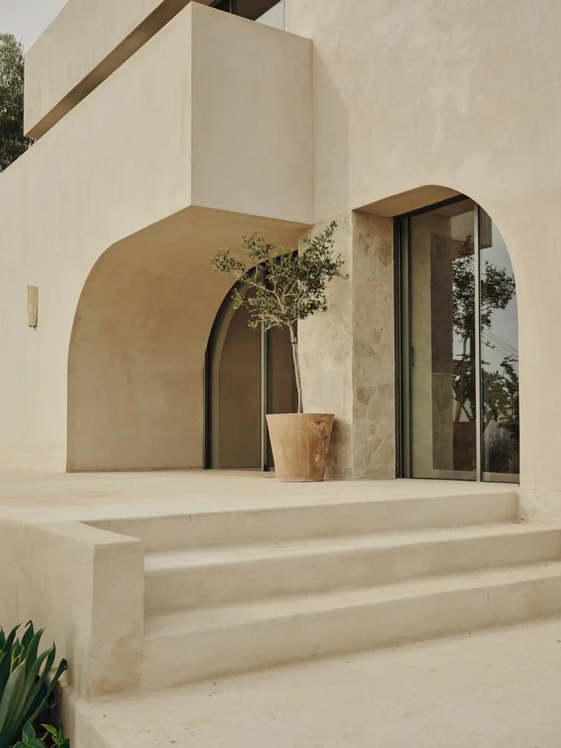 Arched passageway and terrace with potted olive tree and glazed doors under an overcast sky