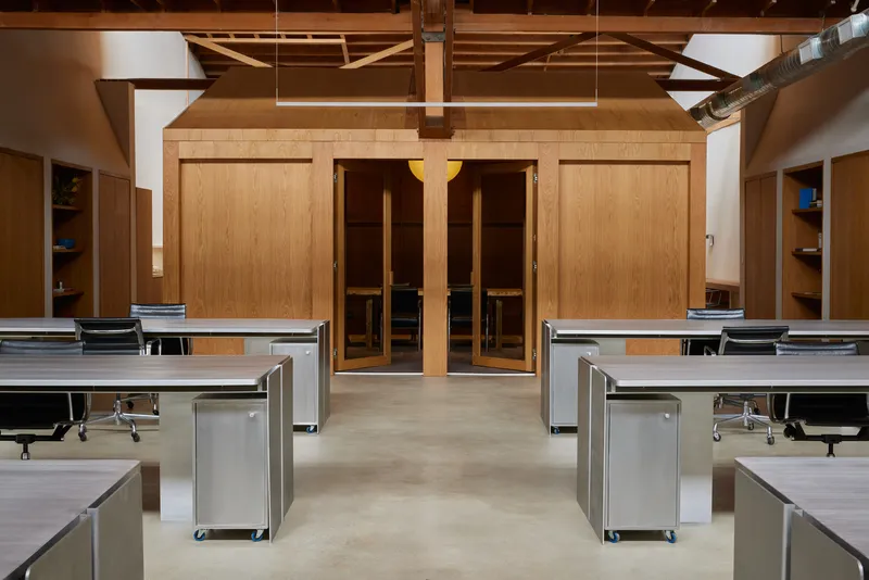 Open office studio with rows of metal desks facing timber-clad sauna doors beneath exposed wood trusses