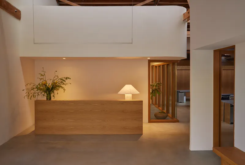Reception desk in pale oak beneath exposed timber beams with potted plants and glimpse of adjacent rooms