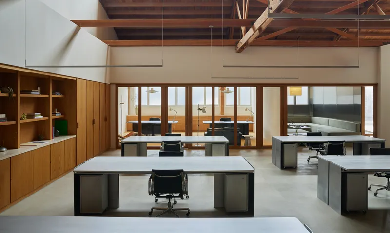 Open office space with exposed timber trusses, wooden millwork wall, and dark metal desks on polished concrete
