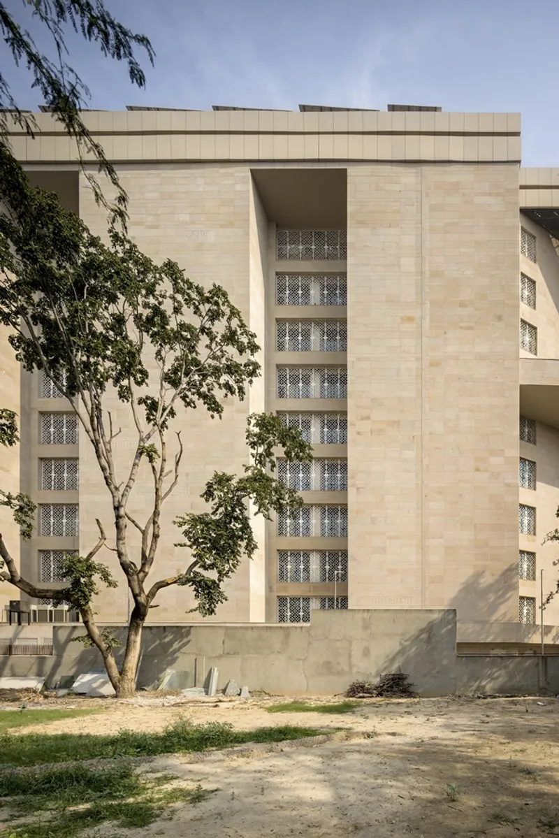Ground-level view of the stone facade with vertical lattice screens framed by a foreground tree