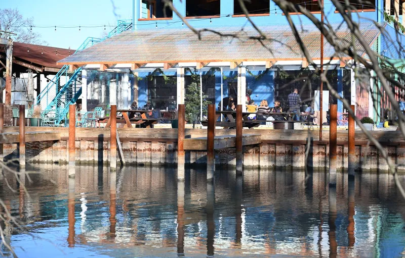 Waterside terrace on timber pilings reflected in calm water with diners seated inside the glazed enclosure