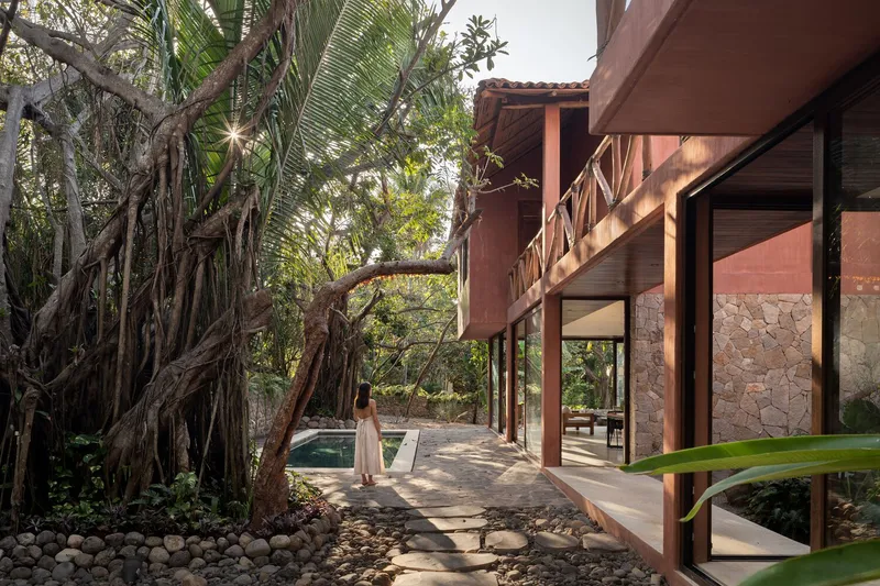 Covered walkway with timber columns and stone walls alongside a pool and banyan tree roots