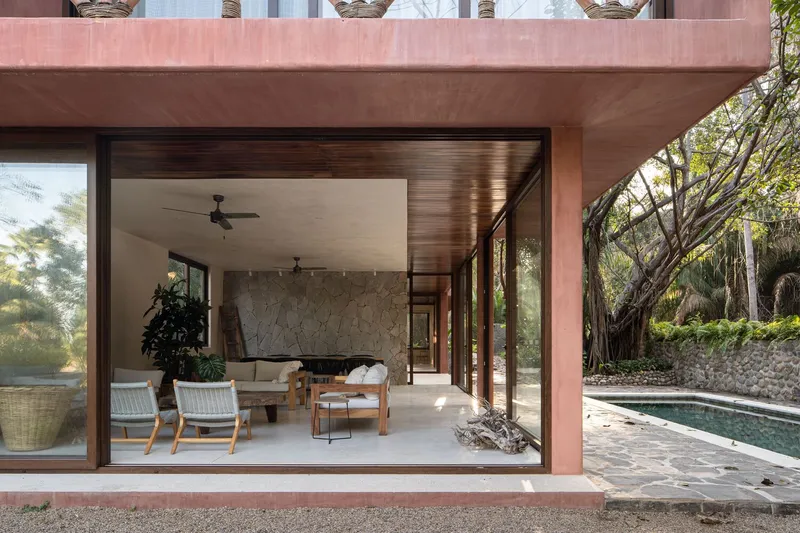 Living room with slatted timber ceiling and stone wall opening to poolside terrace in afternoon light