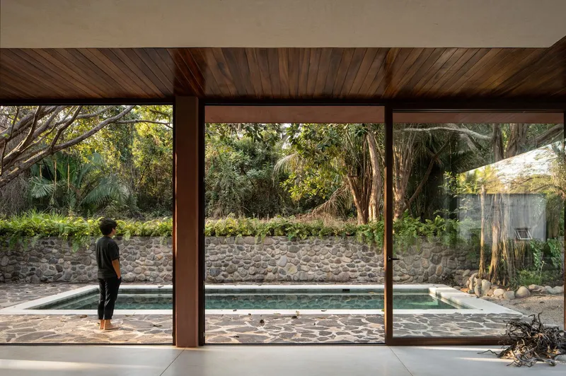 Floor-to-ceiling glass wall framing the pool and cobblestone retaining wall with a figure standing inside