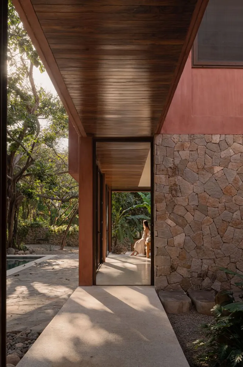 Covered walkway with horizontal wood ceiling and irregular stone wall opening to a garden courtyard with dappled sunlight