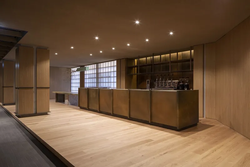 Bar counter clad in brass panels beneath gridded translucent window and timber display shelving
