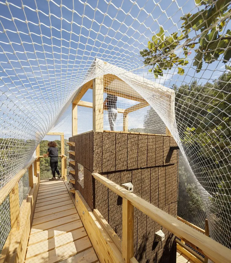 Interior view along mesh-enclosed walkway with timber posts and cork-clad walls under blue sky