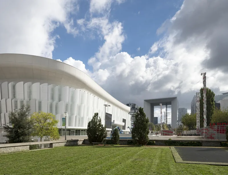 Curved facade with vertical translucent fins across from a lawn and plaza under dramatic clouds