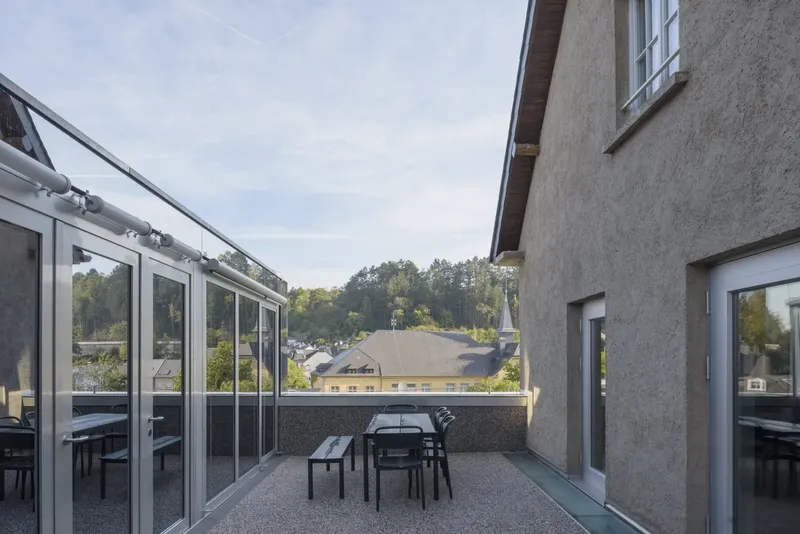 Rooftop terrace view between stucco volume and glazed pavilion with forested hillside beyond