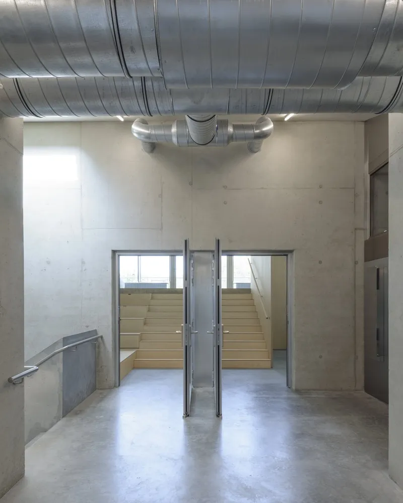 Concrete entrance hall with exposed ventilation ducts and glazed doors opening to timber bleacher seating
