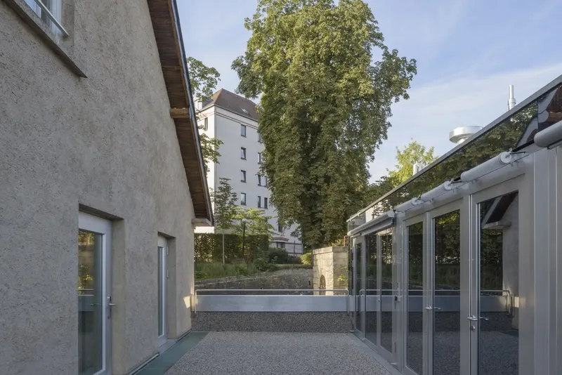 Gravel courtyard flanked by a textured stucco wall and a glazed pavilion under mature trees