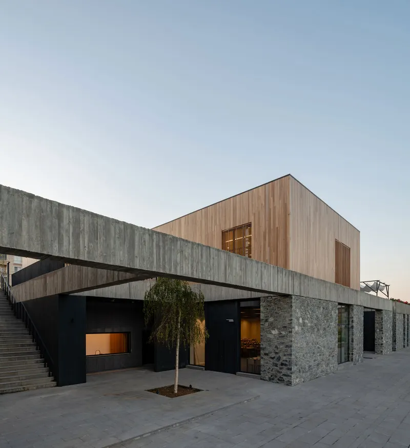 Ground level plaza with stone walls, concrete steps, and timber volume above at dusk